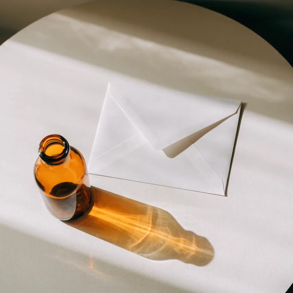Correspondence still life: a plain envelope and an unmarked amber glass bottle on pale board stock under morning window light